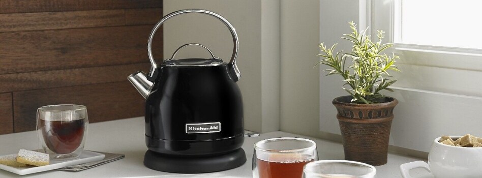A KitchenAid Electric Kettle on a counter surrounded by cups of tea and a potted plant. 